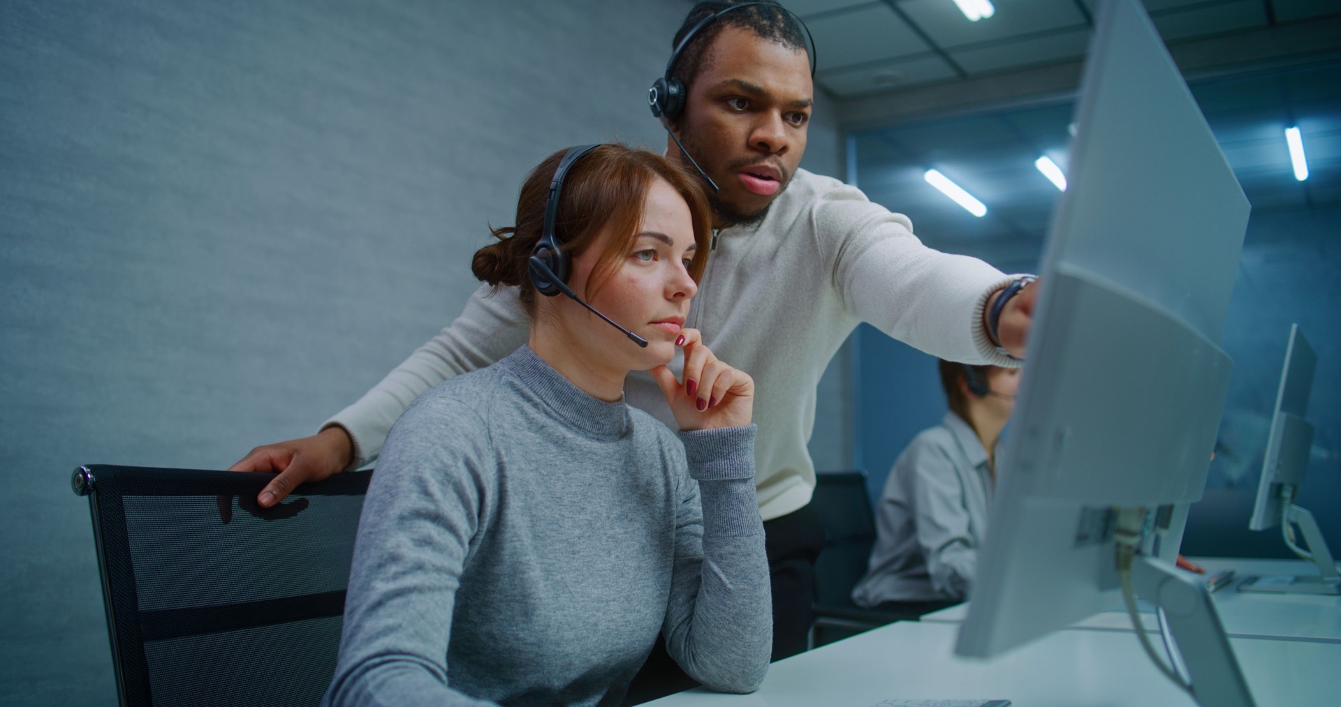 Two multiethnic hotline operators in headsets talk monitoring data on computer