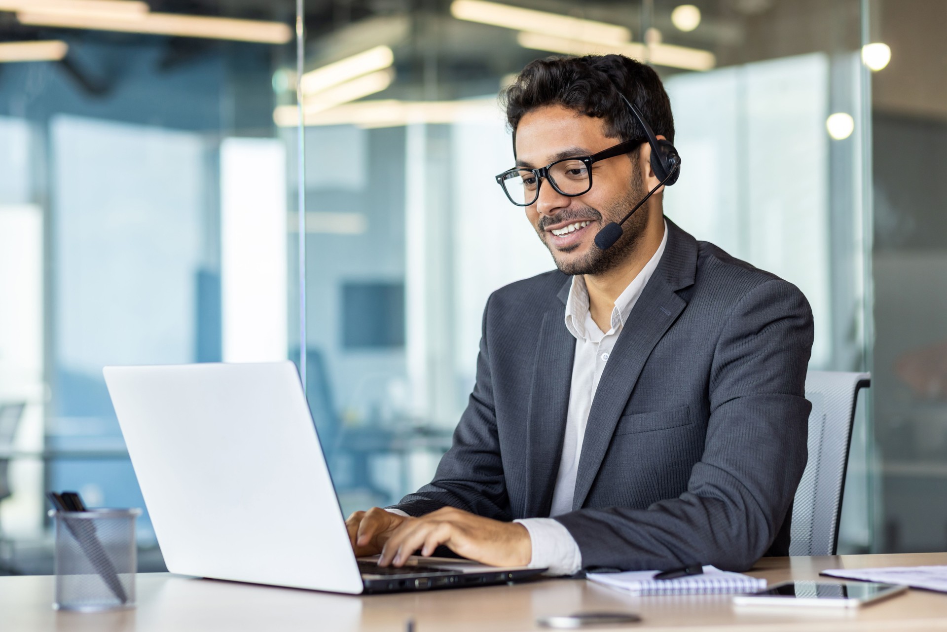 Young successful man at workplace with headset phone typing on laptop keyboard, customer support worker for online buyers inside office, hispanic helping and consulting customers